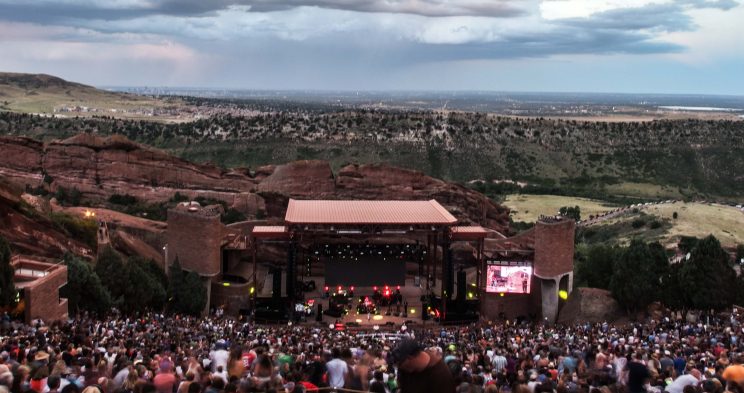 Image: photo of an outdoor amphitheater shot from the back row with the audience between the camera and the stage, with the foothills of Colorado in the background.