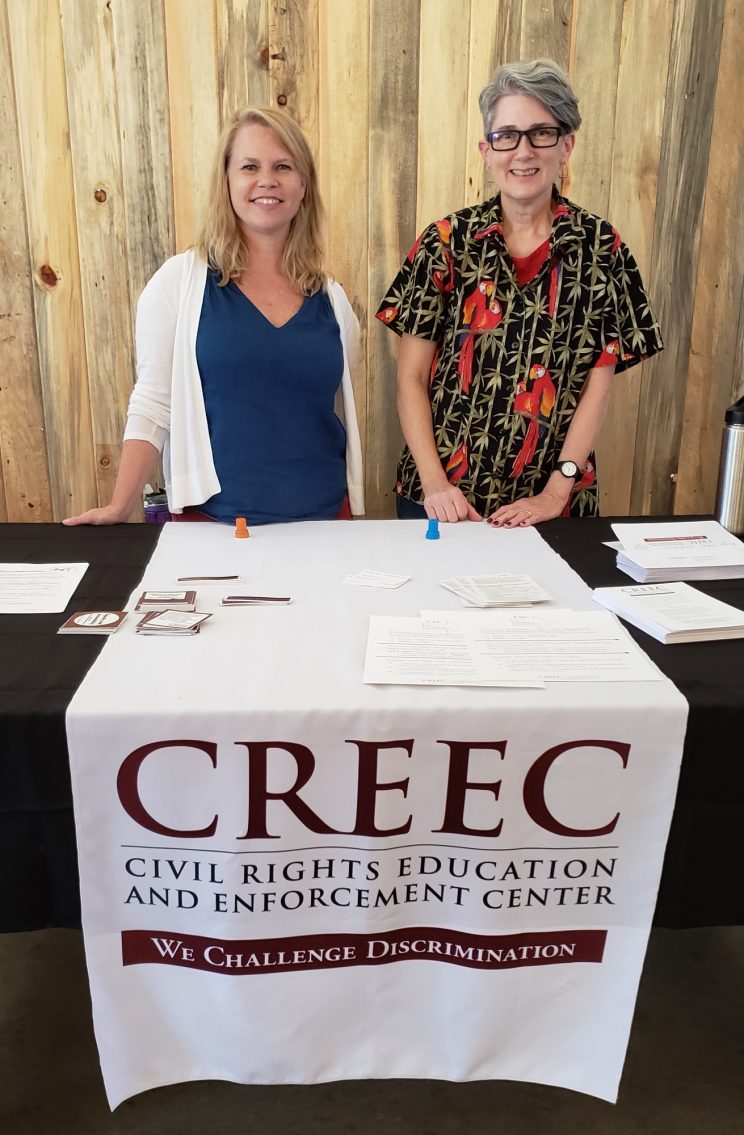 Two white women. Amy is on the right in a parrot-themed Hawaiian shirt; she has short salt & pepper hair and glasses. CREEC’s interpreter is on the left in a dark blue shirt and white sweater; she has shoulder-length blond hair. They are standing behind a table with a banner that reads “CREEC Civil Rights Education and Enforcement Center. We challenge discrimination.” 