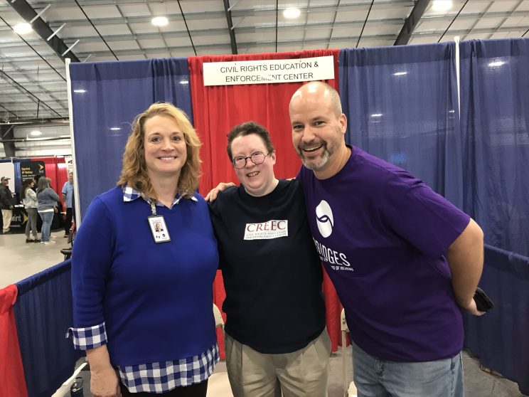Two white women and one white man. Martie is in the middle wearing a blue shirt with the CREEC logo. Our interpreter for the event, Beth Pilkington, is on the left in a blue shirt. On the right in a purple shirt is Mike Helms, VP of Adult Education and Outreach at Bridges in Nashville. Behind the trio is the CREEC booth with red and blue curtains and a sign that says Civil Rights Education and Enforcement Center.