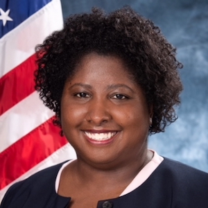 A photograph of Rabia Belt. Rabia is a Black woman wearing a dark professional shirt. She is smiling and is in front of a blue backdrop with an American flag.