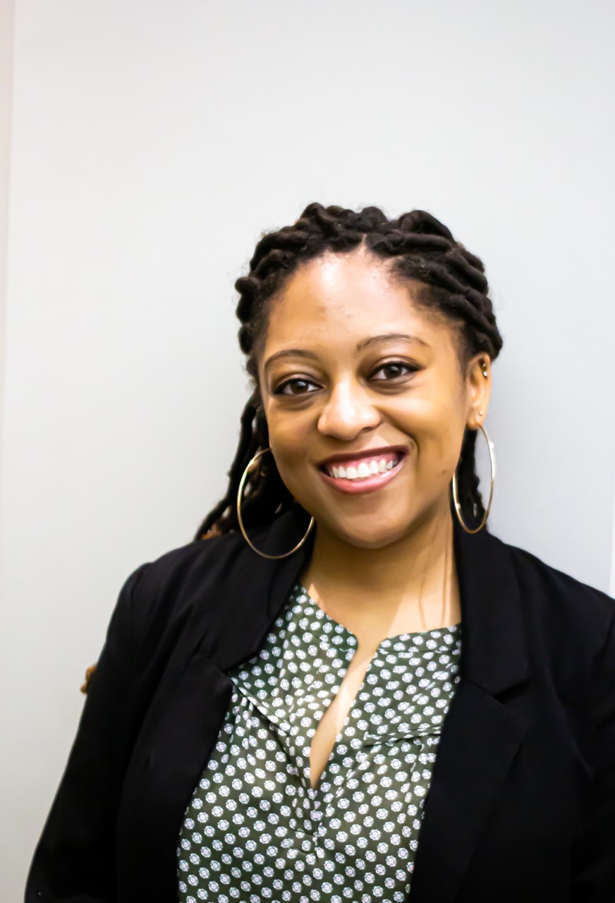 Photograph of Toni Cross. Toni is a Black woman wearing a black blazer, a dark green and white shirt, and metallic hoop earrings. She is smiling. The background is a white wall.