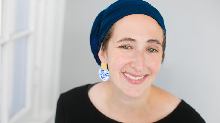 A photo of Lauren, a woman with light skin and hazel eyes who is smiling. She’s wearing a blue tichel (head scarf), a black shirt, and circular gold, blue, and white earrings.