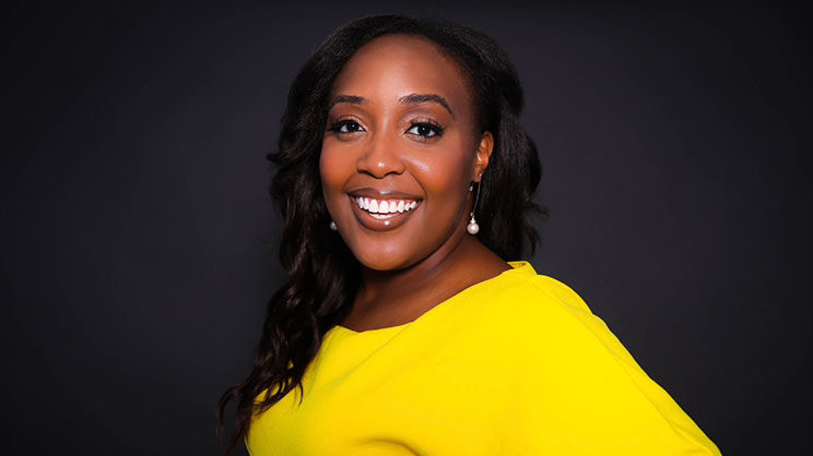 Photo of Aviance Brown, a Black woman wearing a bright yellow dress with long loosely curled dark brown hair, dangling pearl earrings and she is smiling in front of a black background.