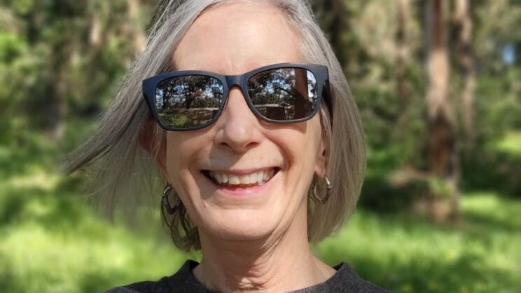 Photo of Cynthia, a white woman wearing sunglasses with medium length gray hair, smiling in a forest with the wind blowing her hair.