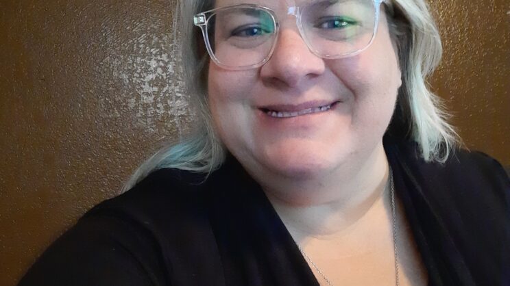 Photo of Sara, a white female with short length white/blonde hair. She's smiling and is wearing clear eyeglasses with black knit sweater and a maroon top. She is also wearing a silver elephant necklace.